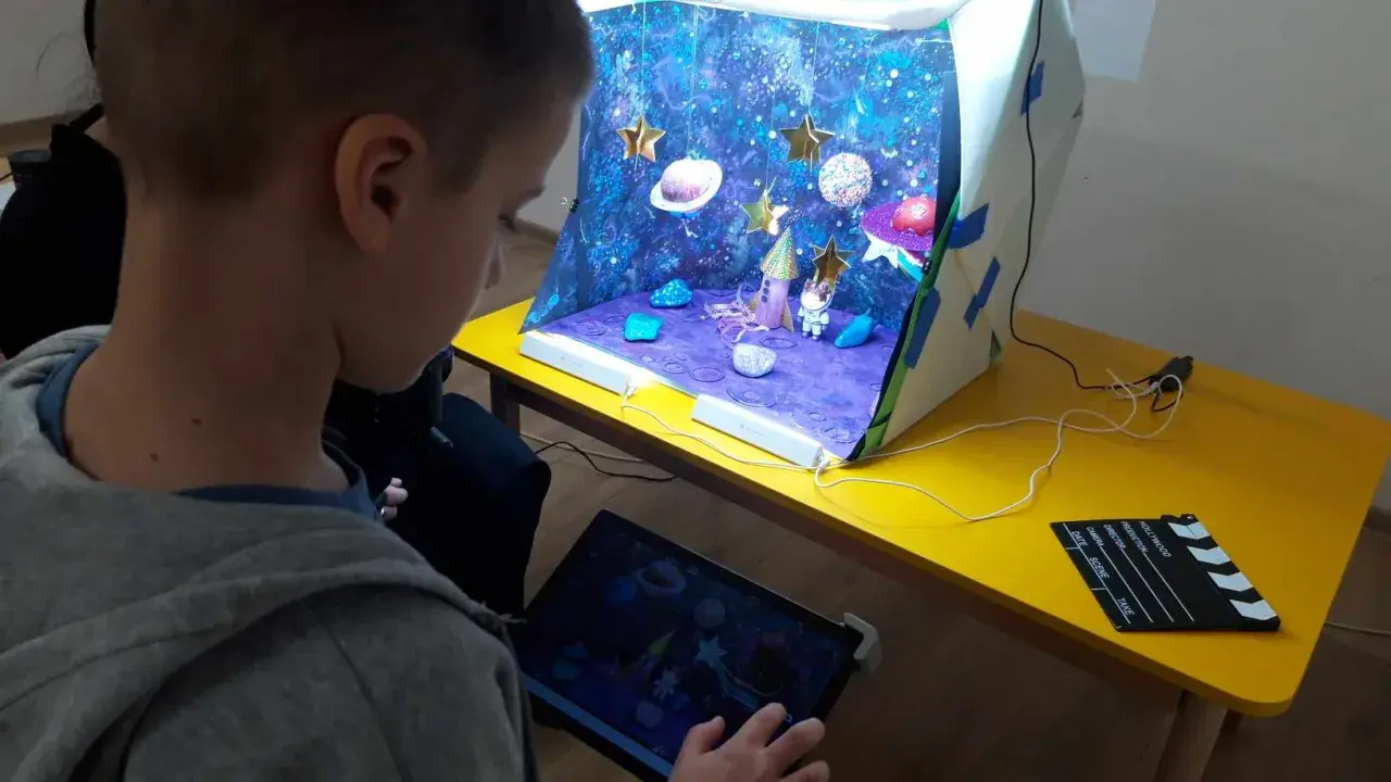 Chłopiec tworzy kosmiczną animację poklatkową. Na stole stoi diorama z rakietą, astronautą i planetami, a obok tablet z podglądem.