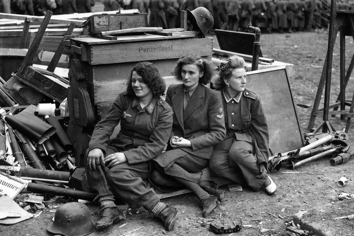 Trzy kobiety w mundurach siedzą obok skrzyń z napisem "Panzerfaust". To poruszająca fotografia wojenna.