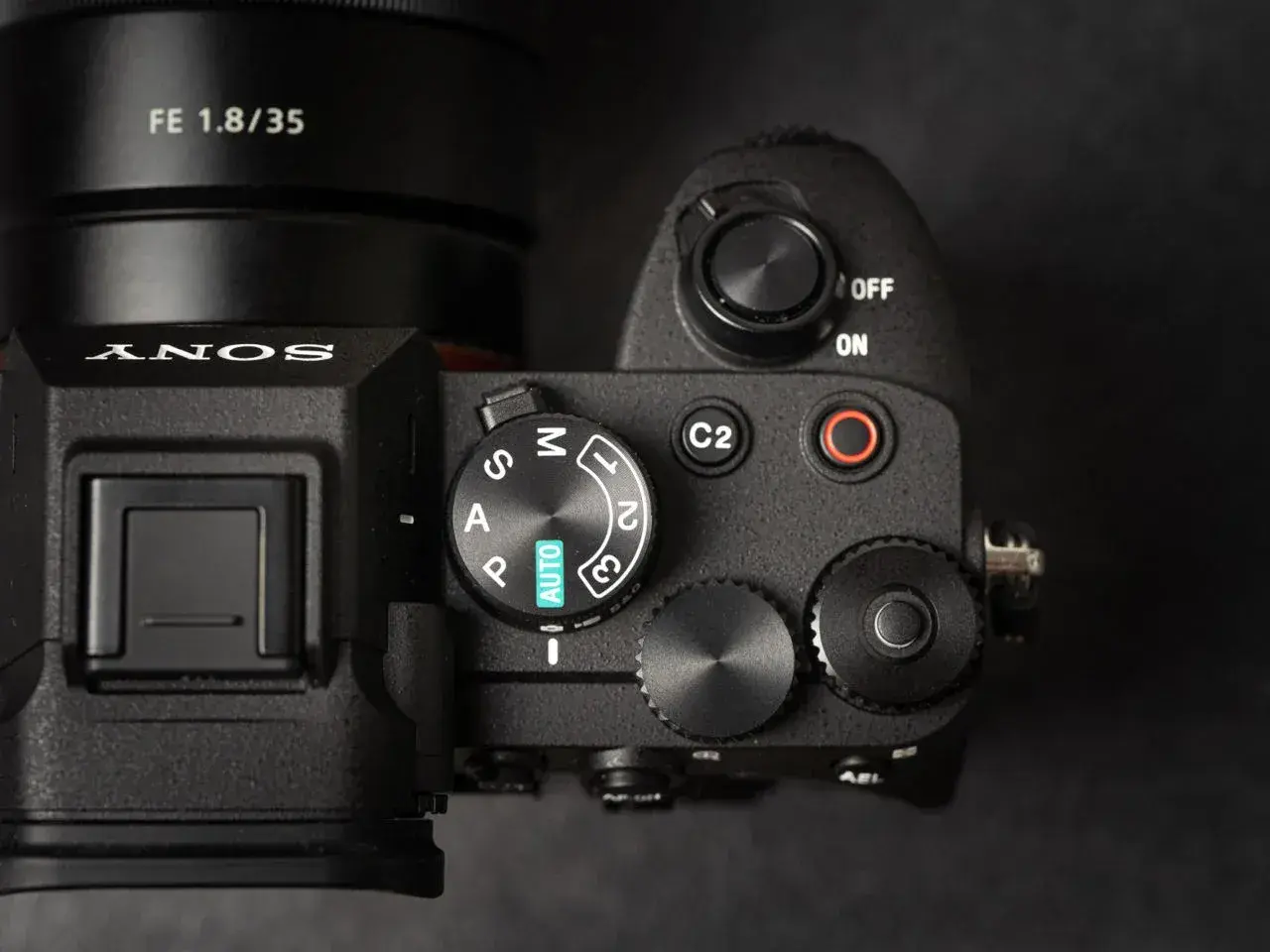 Zbliżenie na górny panel aparatu Sony A7 IV, gotowego na premierę. Widoczny obiektyw FE 1.8/35 i pokrętło trybów.