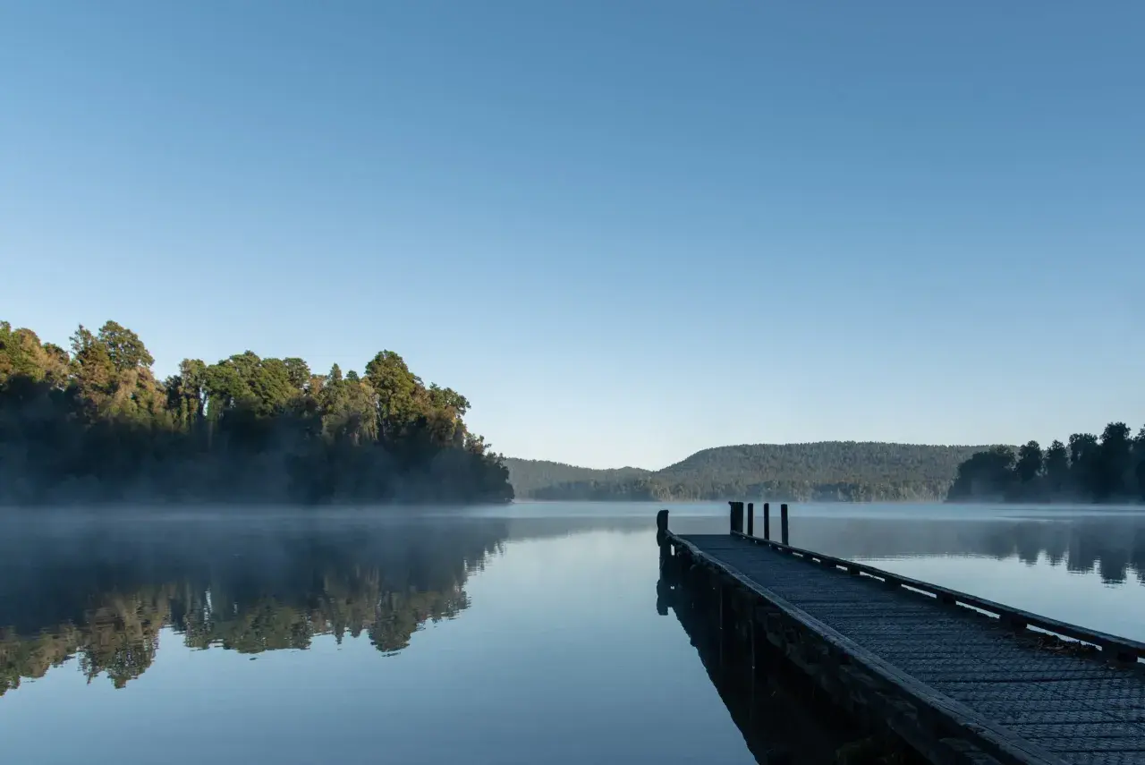 Spokojny wschód słońca nad jeziorem, mgła unosi się nad wodą, a drewniane molo zaprasza do spaceru.
