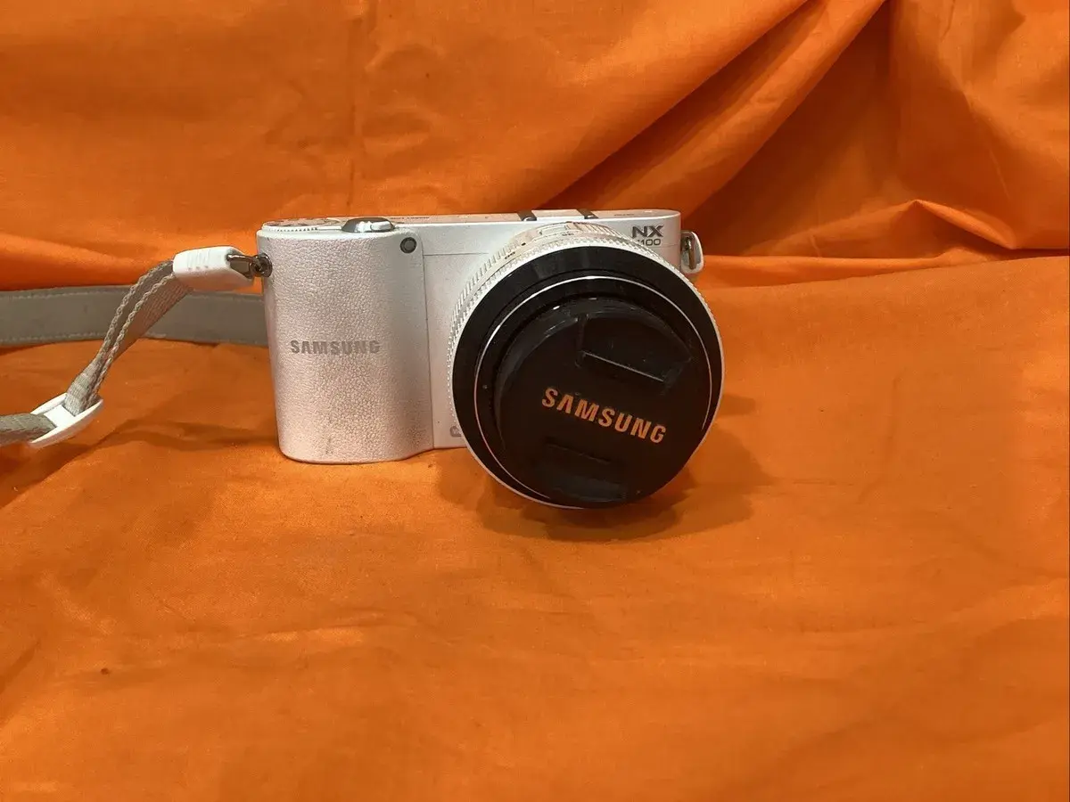 Biały aparat Samsung NX1100 z obiektywem i dekielkiem z logo Samsung, na pomarańczowym tle.
