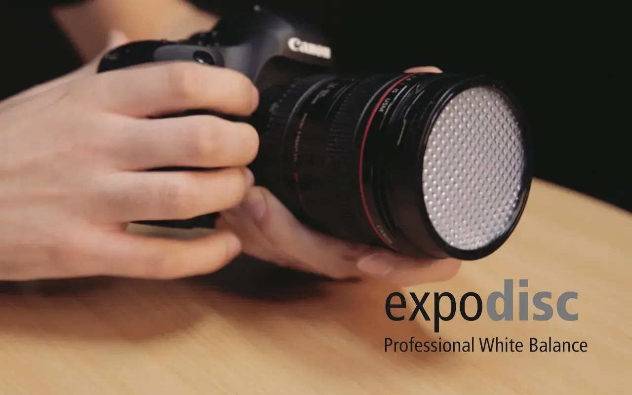 Dłonie trzymają aparat z obiektywem, na którym zamontowano biały dyfuzor. Obok widoczne logo "expodisc Professional White Balance".