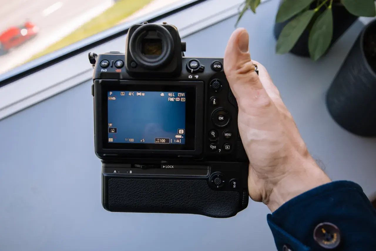 Dłoń trzyma aparat Nikon Z8, testując jego funkcje. Na ekranie widać ustawienia, a w tle rozmyty czerwony samochód.