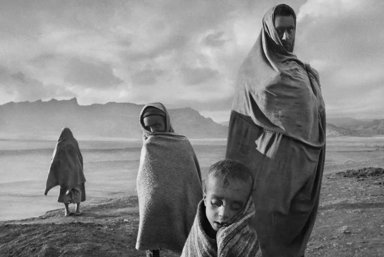 Dzieci i dorosły owinięci w tkaniny, w surowym krajobrazie. Wzrok przyciąga spojrzenie jednego z nich, uchwycone przez Sebastiao Salgado.