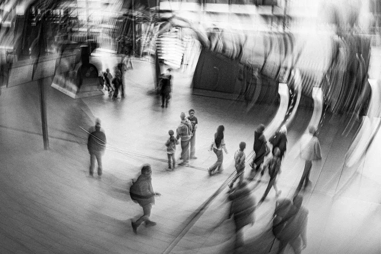 Rozmyta fotografia miejska ukazuje ruchliwą stację kolejową, gdzie sylwetki ludzi zlewają się w dynamicznym tańcu.
