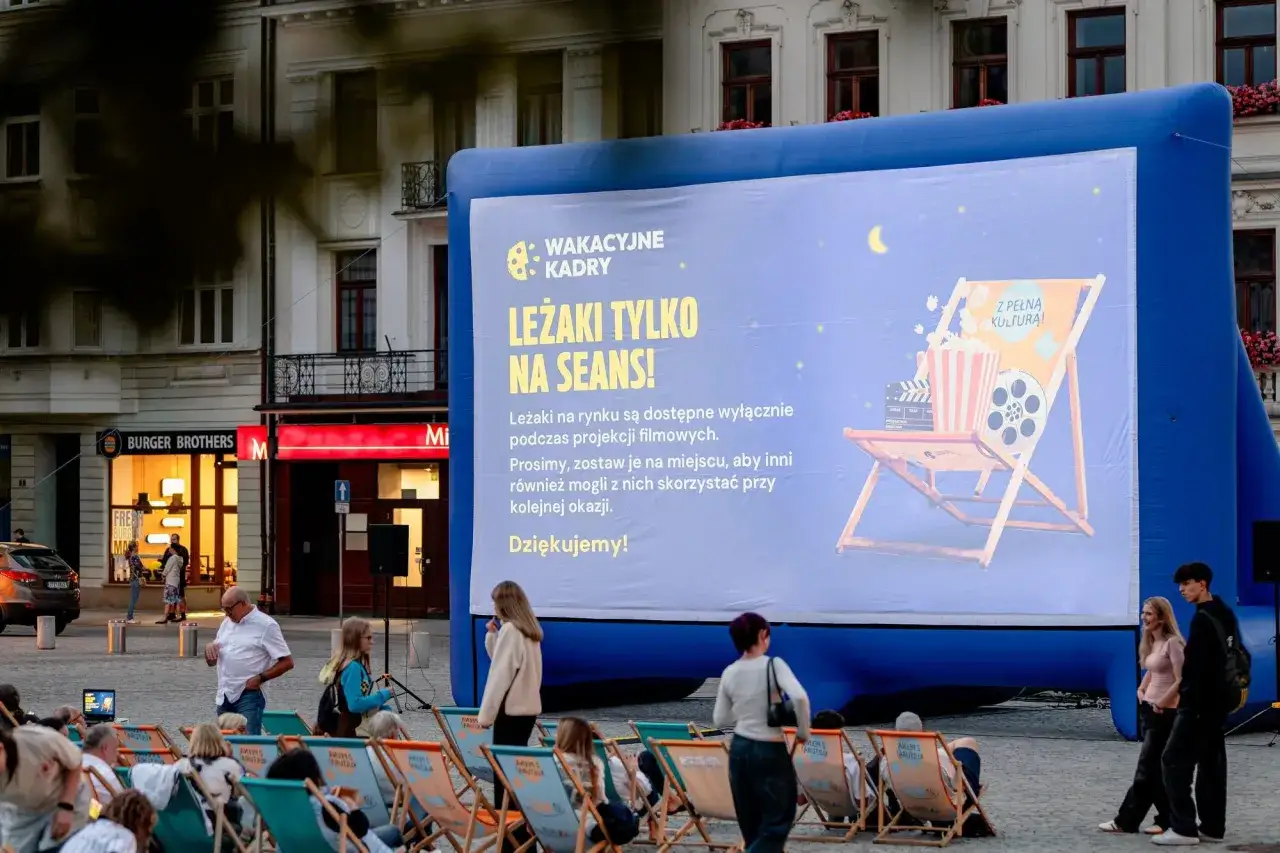 Letnie kino plenerowe. Ludzie siedzą na leżakach przed wielkim ekranem, gdzie wyświetlane są napisy 