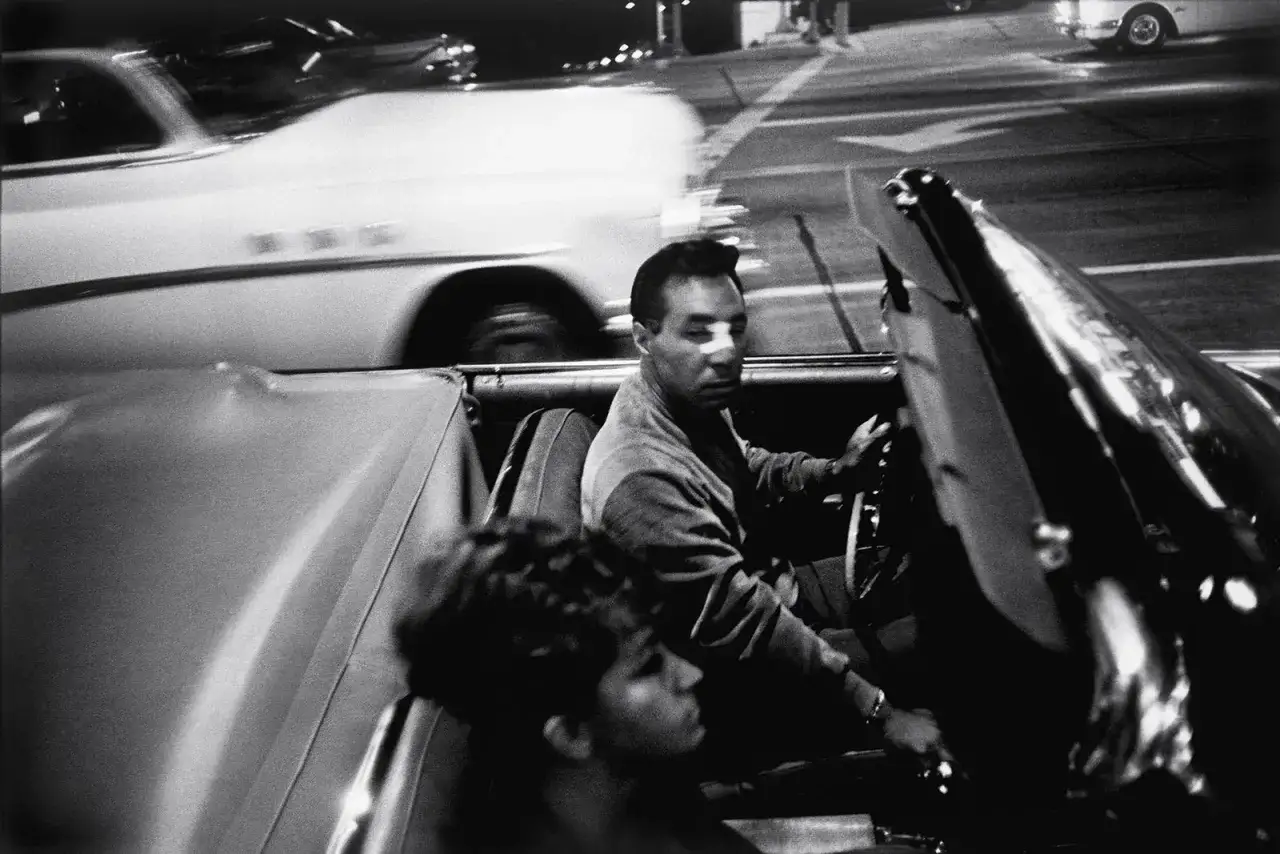 Mężczyzna za kierownicą kabrioletu, uchwycony przez garry winogrand, w ruchu ulicznym. Obok siedzi kobieta.