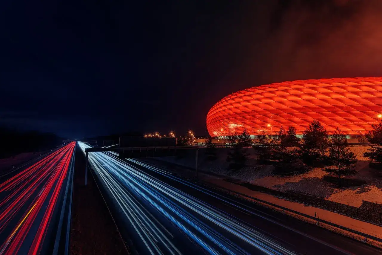 Nocna autostrada z czerwonymi i niebieskimi smugami świateł samochodowych, obok stadionu oświetlonego na czerwono. Efektowne techniki fotograficzne.