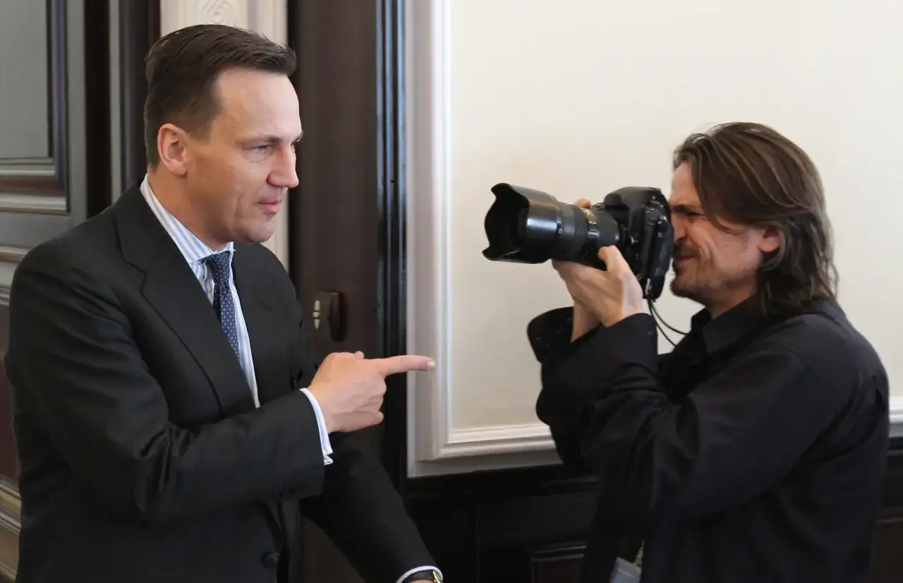 Adam Chełstowski w garniturze wskazuje palcem na fotografa z aparatem.