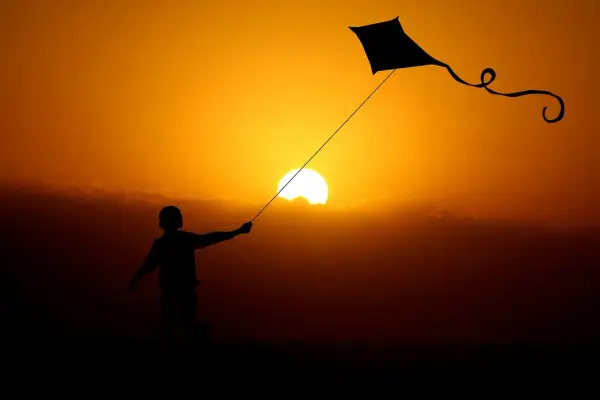 Dziecko puszcza latawiec na tle zachodzącego słońca. Ciepłe światło w fotografii podkreśla sylwetki i tworzy magiczną atmosferę.
